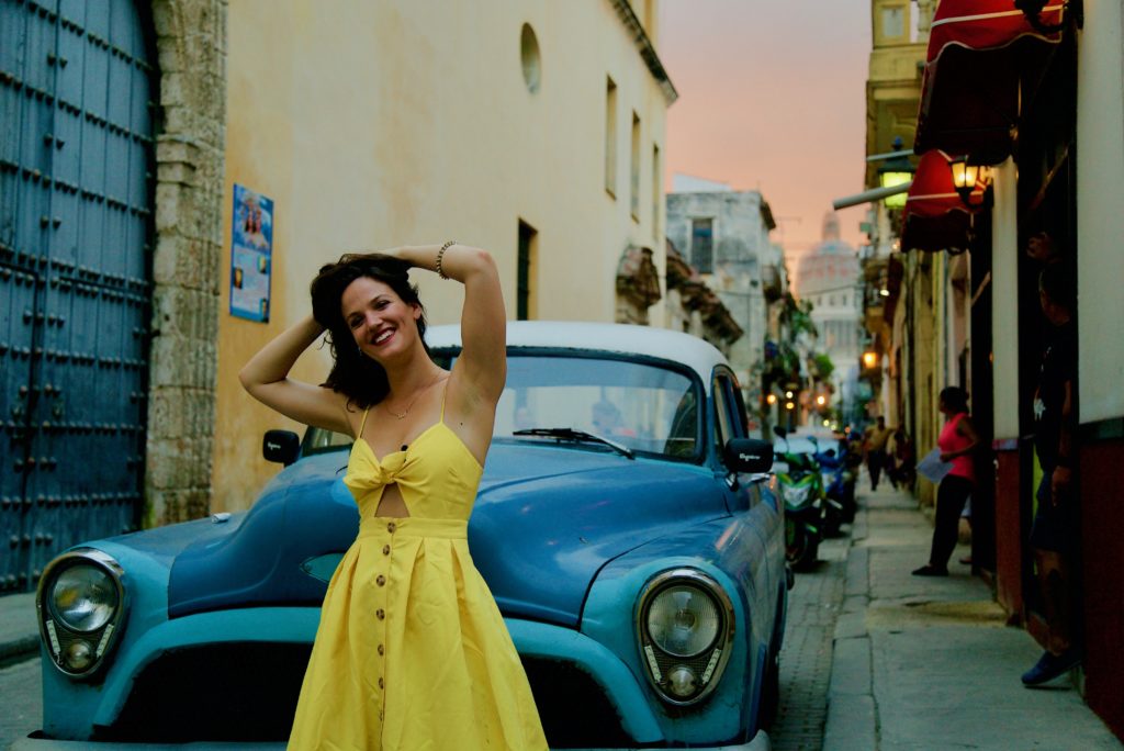 Femme en robe jaune souriant devant une voiture bleue à Cuba